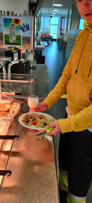 Opiskelija ruokalan noutopöydän äärellä kädessään ruokalautanen ja maitolasi.