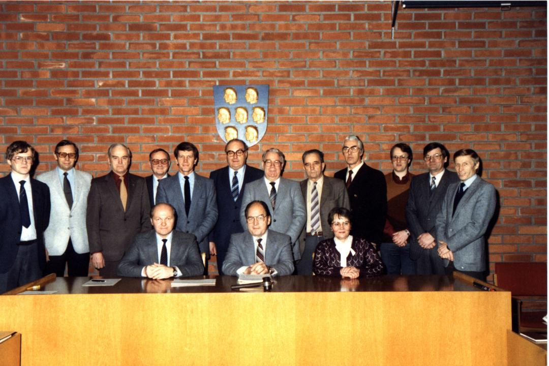 Nurmijärven kunnan päättäjiä Nurmijärven kunnantalon valtuustosalissa, 1984. Kuvassa vasemmalta kunnansihteeri Jukka Anttila, Jouko Narinen, Esa Toivonen, Paavo Taanila, Timo Hyvämäki, Veijo Lehtoruusu, Jorma Jokinen, Matti Mattila, Martti Kostiainen, Hannu Toikkanen, Pertti Lehti ja kunnanjohtaja Toivo Seppälä. Istumassa pöydän takana vasemmalta Esko Moilanen, Mauno Tenhovirta ja Eila Lindholm. Seinällä Nurmijärven kunnan vaakuna.