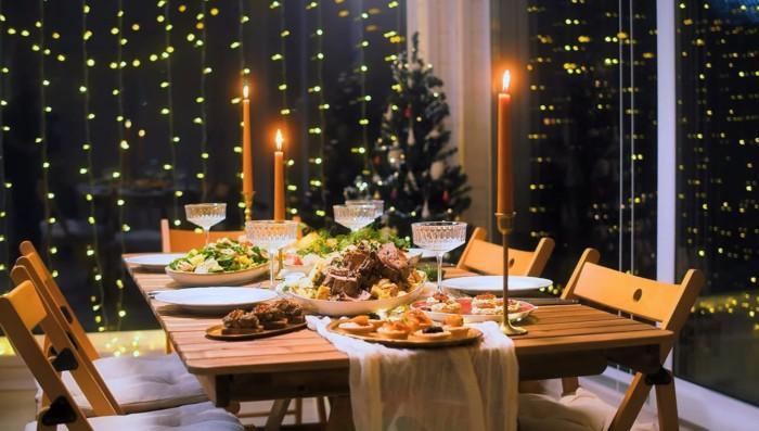 Ruokaa ja juhla-astiat katettuna pöydälle, jossa kynttilät palaa. Taustalla jouluvaloja.