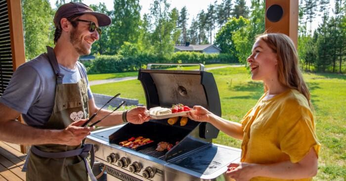 Mies ja nainen grilllin äärellä. Mies ojentaa naiselle lautasta, jossa grilliruokaa.