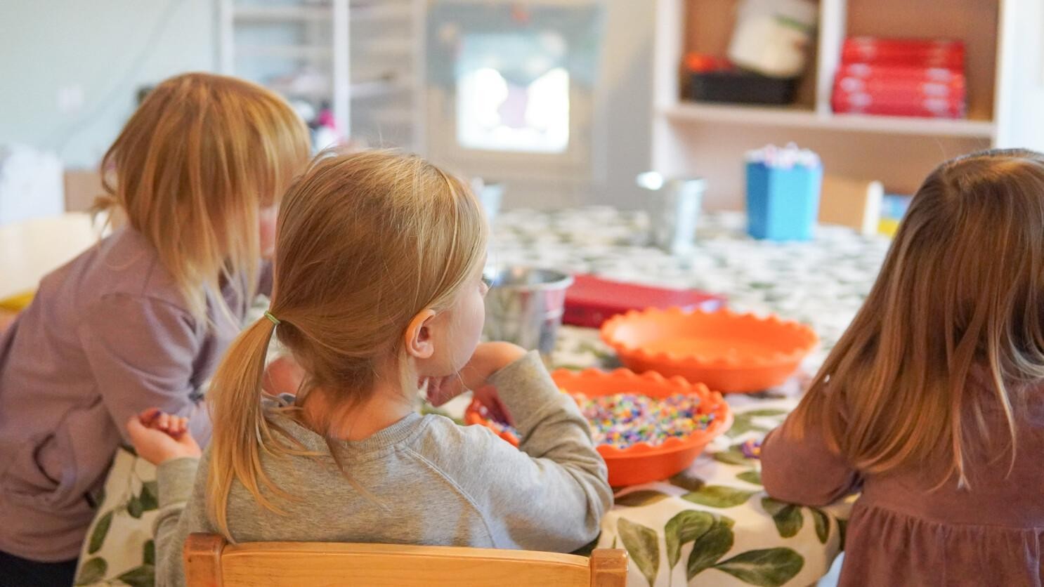 Kolme eskari-ikäistä lasta kuvattuna takaapäin. Istuvat pöydän ääressä askartelemassa, katse suunnattuna oikealle.